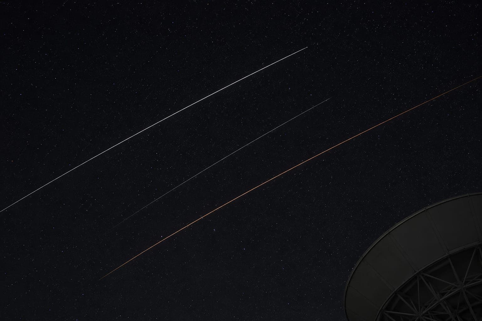 Long-exposure photograph of low-earth-orbit satellites streaking across a starfield, with the silhouette of a ground-station antenna in the lower right; one streak glows warm orange