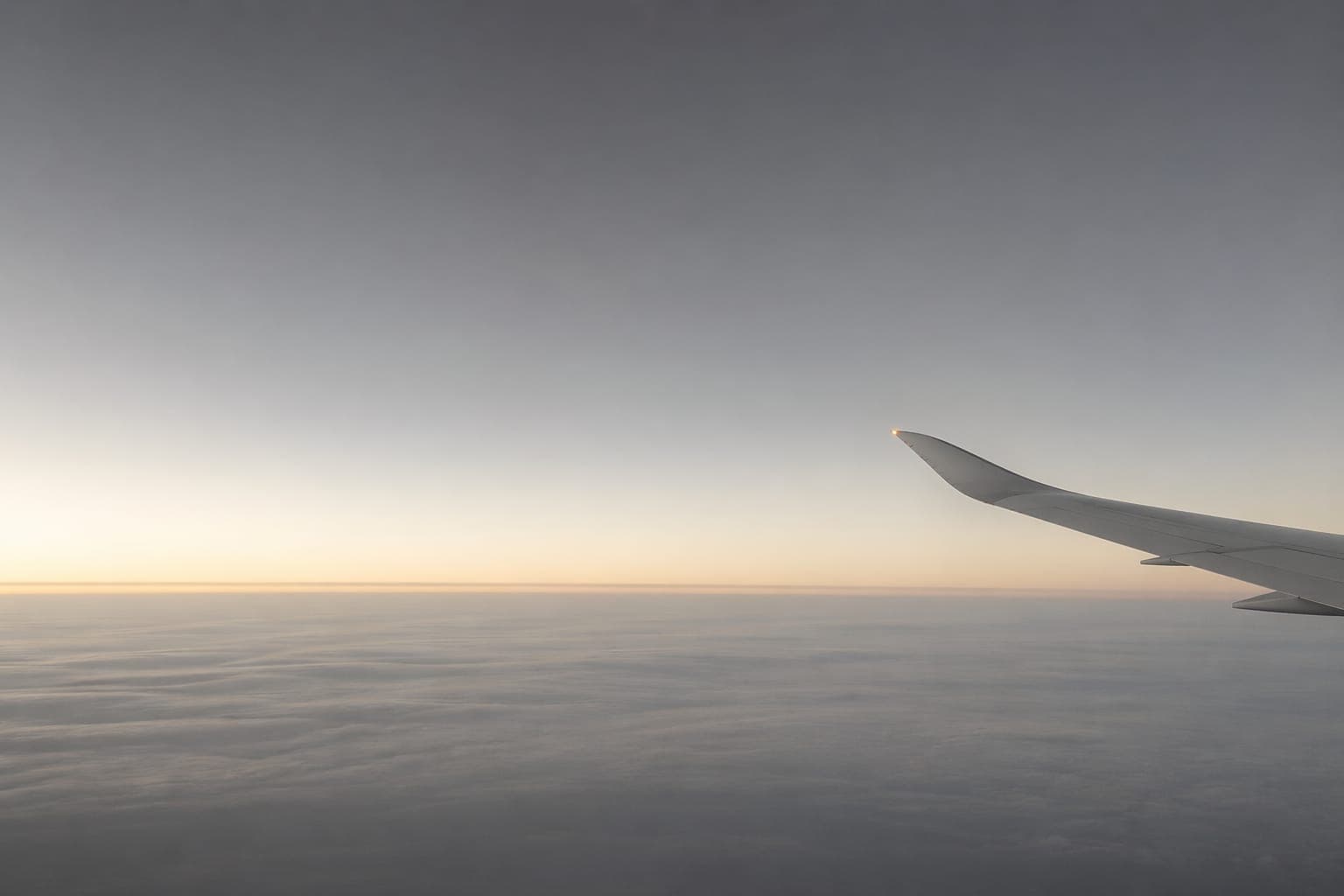 A FlyLo aircraft wing seen from a window seat at dusk, the horizon a pale peach line, soft grey clouds below — the quiet aircraft in flight